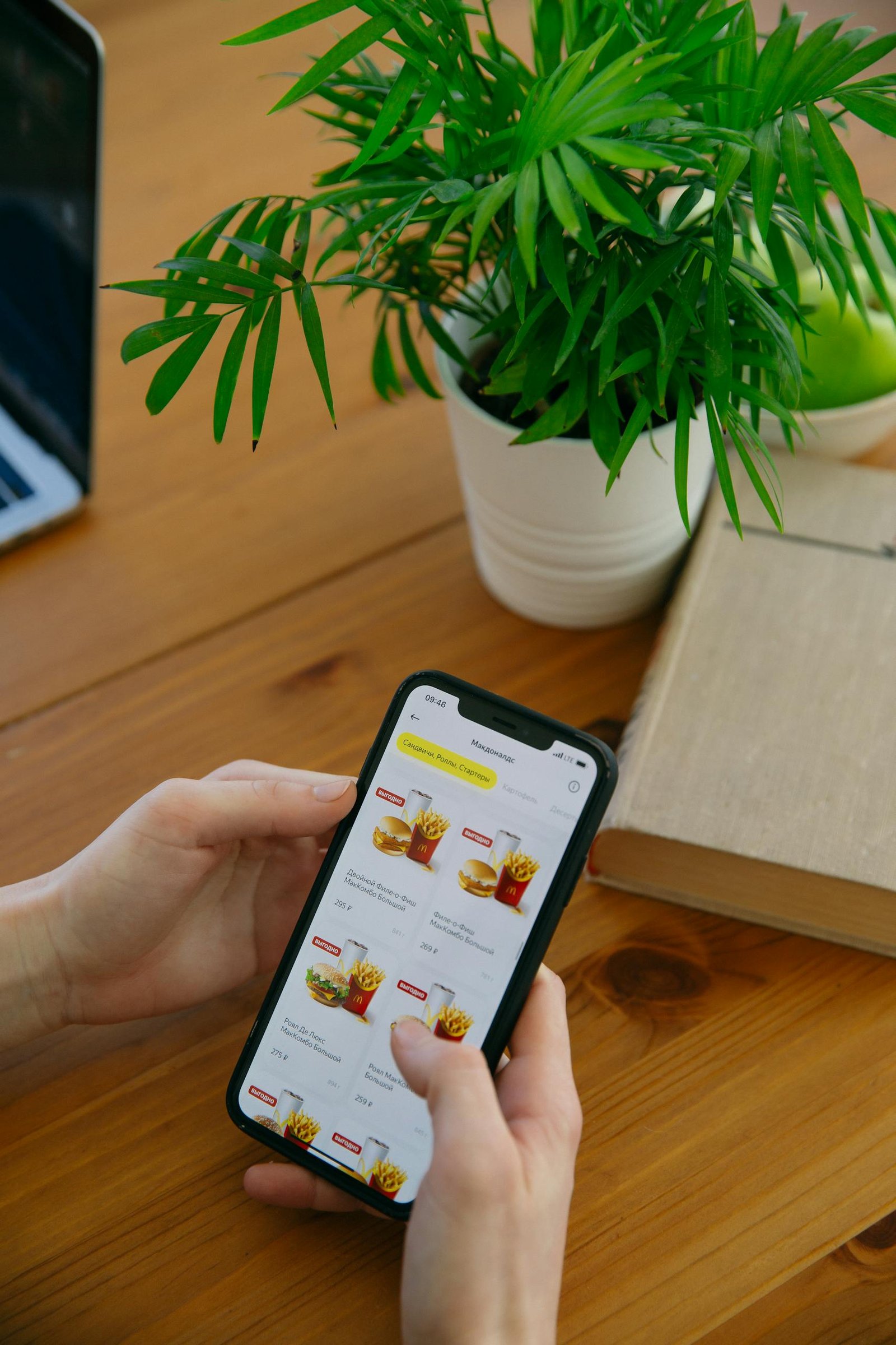 Hands using smartphone to browse food delivery app next to a potted plant.